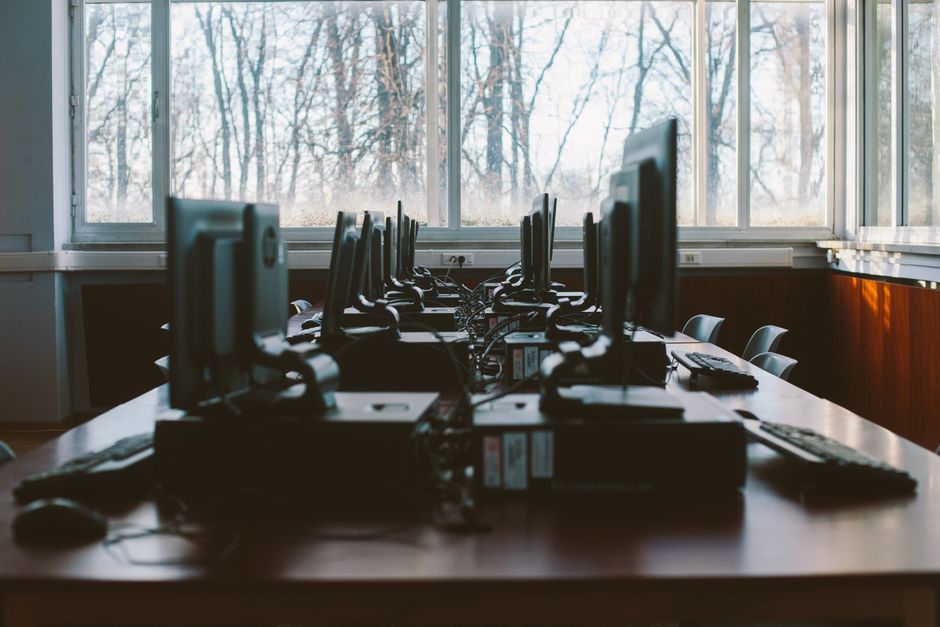 Sala de informática com computadores em ambiente de treinamento ou estudo, com janelas que deixam passar luz natural e revealing uma paisagem de árvores ao fundo.