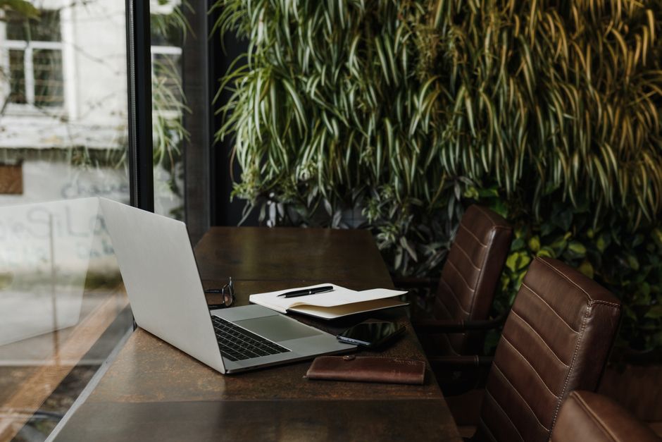 Escritório aconchegante com laptops e materiais de trabalho ao lado de uma janela, rodeado por plantas verdes, ideal para artigos sobre trabalho remoto ou ambientes de trabalho inspiradores.