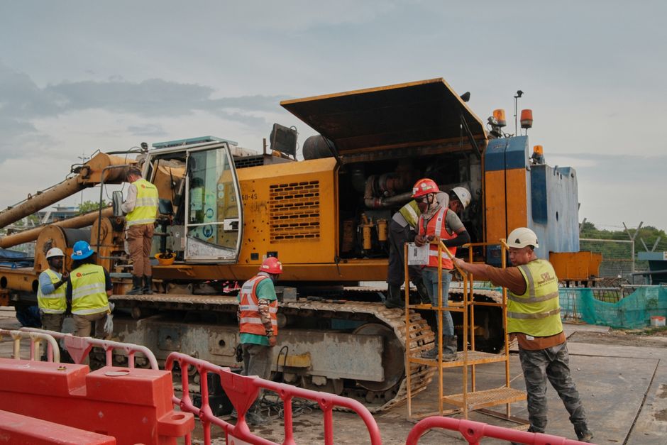 Equipe de trabalhadores da construção civil operando equipamento pesado em um canteiro de obras ao ar livre.