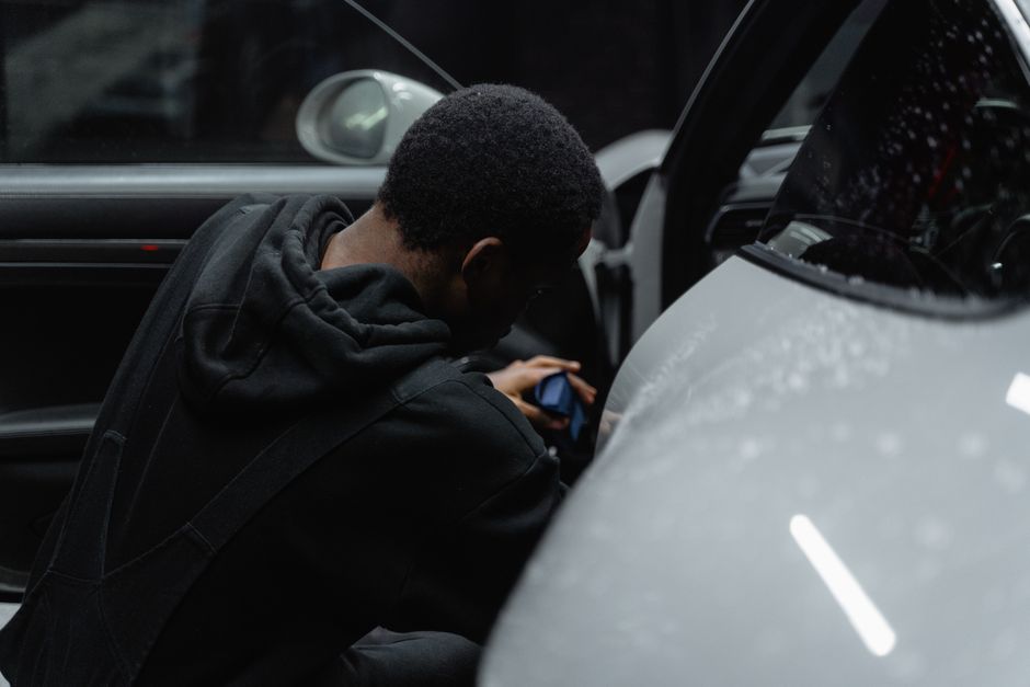 Homem sentado ao lado de um carro, revisando documentos ou verificando informações, em um ambiente externo com foco na preparação ou inspeção automotiva.