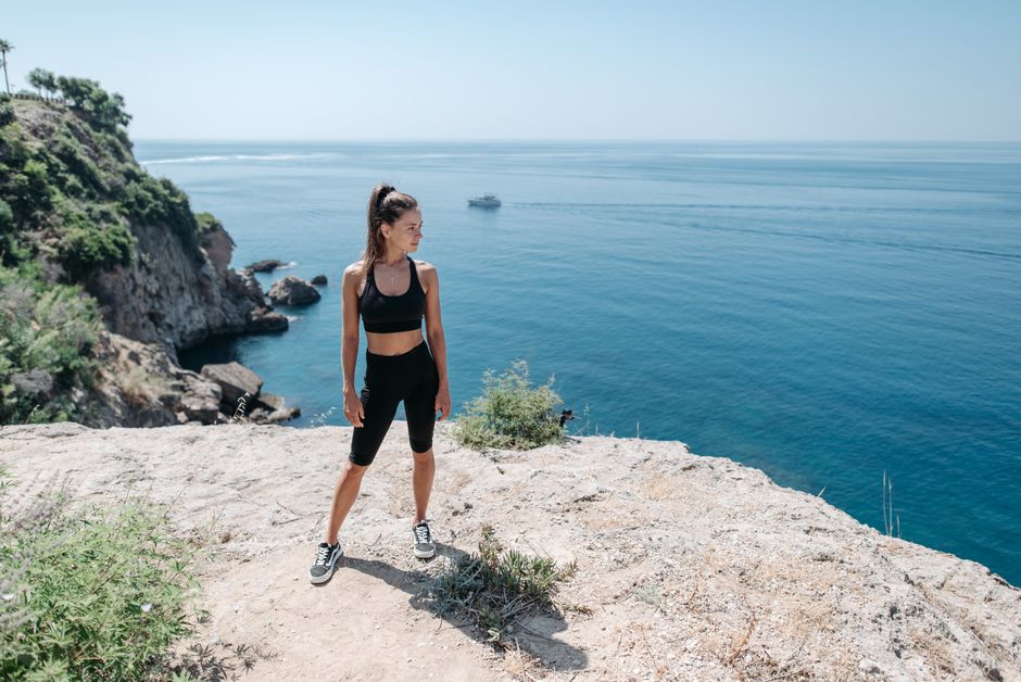 Mulher praticando caminhada ou corrida em trilha com vista para o oceano e clima de verão ao fundo.