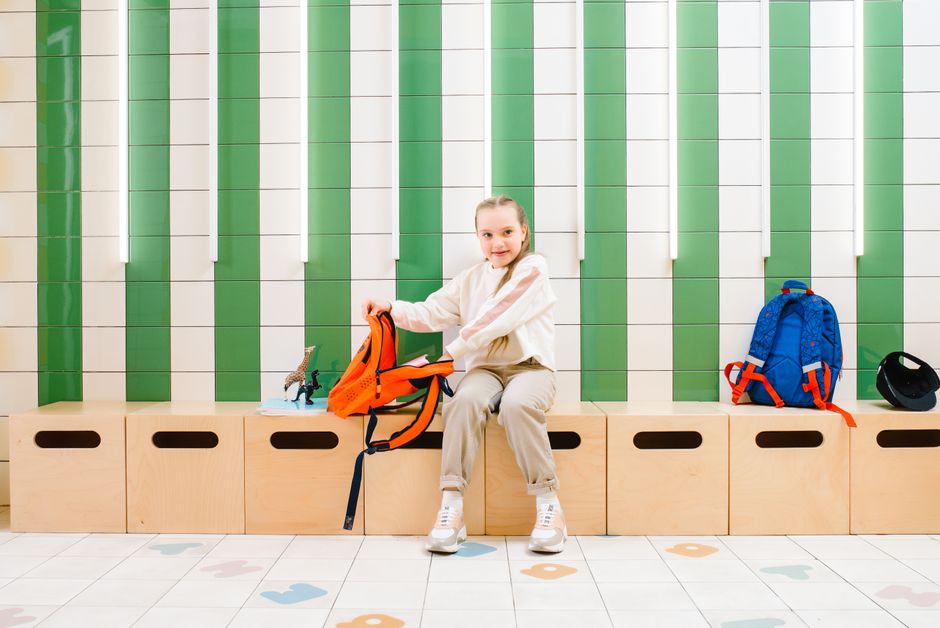 Menina sorridente sentada em banco de espera na escola, preparando-se para a aula com mochila ao lado.