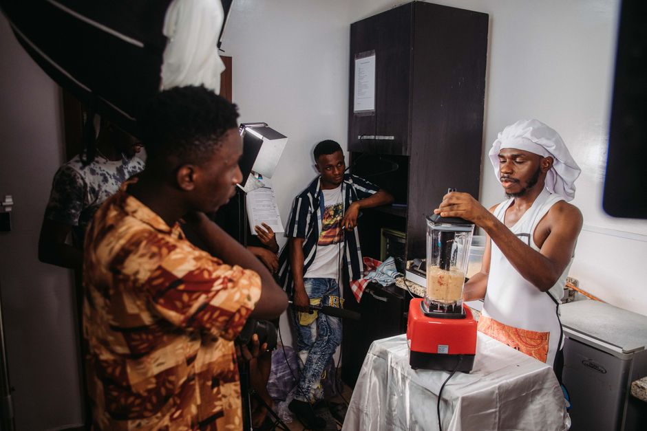 Homem participando de uma aula de culinária em ambiente interno, com outros presentes observando atentamente junto ao participante, que está preparando uma receita com um liquidificador.