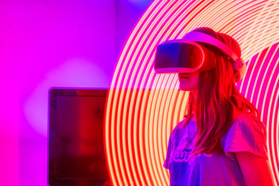 Imagem de uma mulher usando óculos de realidade virtual em um ambiente com iluminação colorida, representando inovação tecnológica e experiências imersivas.