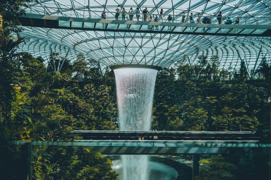 Interior de um ambiente futurista com uma cascata artificial integrada a um espaço de convivência com muitas plantas e pessoas caminhando na passarela superior.