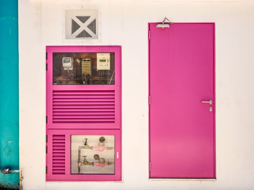 Imagem de uma parede com uma porta e uma caixa de energia, ambas pintadas em rosa vibrante, criando um contraste marcante na composição.