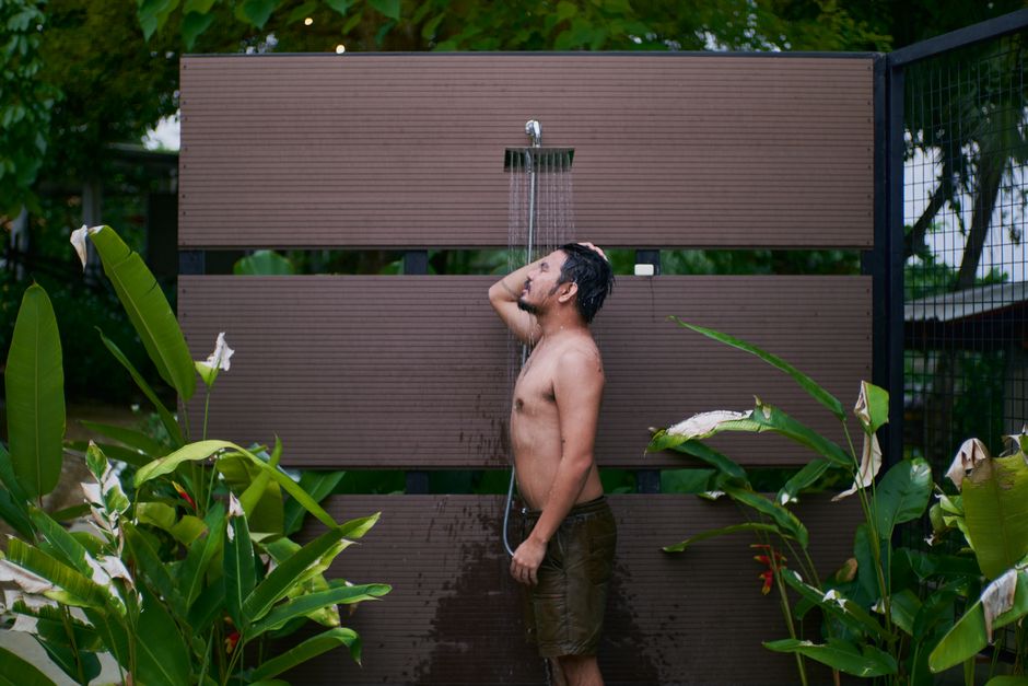 Homem tomando banho sob ducha ao ar livre, rodeado de vegetação tropical, em um ambiente de lazer ou relaxamento ao ar livre.