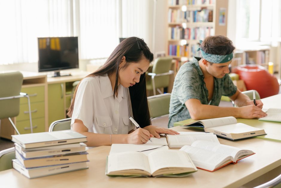 Estudantes concentrados estudando em uma biblioteca bem iluminada, com livros abertos na mesa.