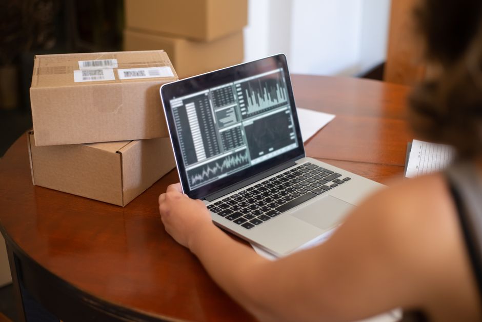 Pessoa analisando dados de vendas ou desempenho de negócios em um laptop ao lado de caixas de entrega, em um ambiente de escritório ou logístico.
