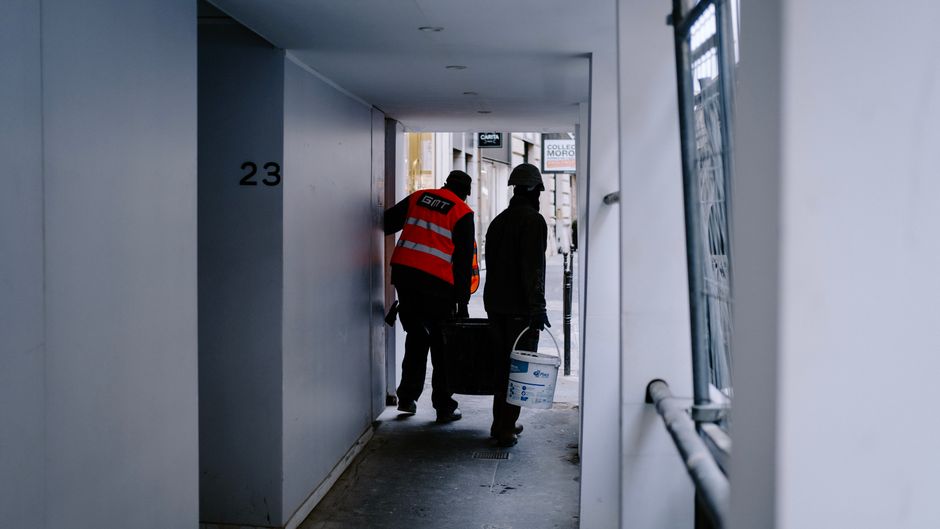 Dois trabalhadores carregando materiais de construção em uma passagem estreita de edifício em uma área urbana, relacionados a reformas ou entregas em obras civis.
