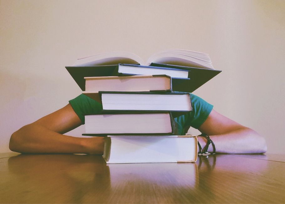 Pessoa com o rosto coberto por pilhas de livros que formam uma cabeça de aparência criativa e divertida, simbolizando estudo ou conhecimento acumulado.