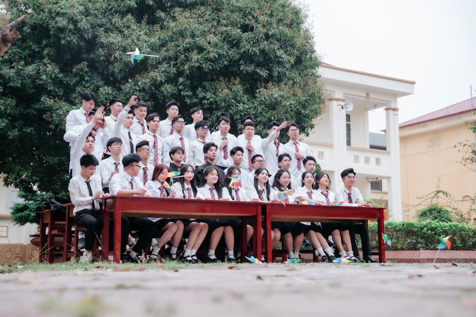 Grupo de estudantes em aula ao ar livre, participando de uma atividade escolar coletiva com bandeiras e aviões de papel, simbolizando colaboração e criatividade na educação.
