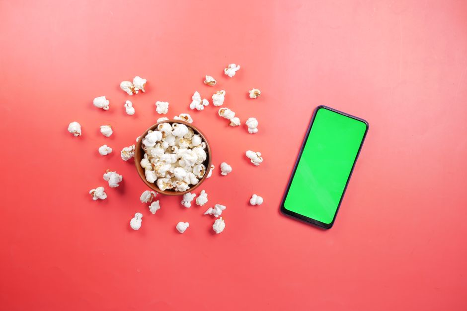 Imagem de uma tigela de pipoca ao lado de um smartphone com tela verde, sobre fundo rosa, ideal para ilustrar distra&ccedil;&otilde;es durante o lazer ou consumo de m&iacute;dia digital.