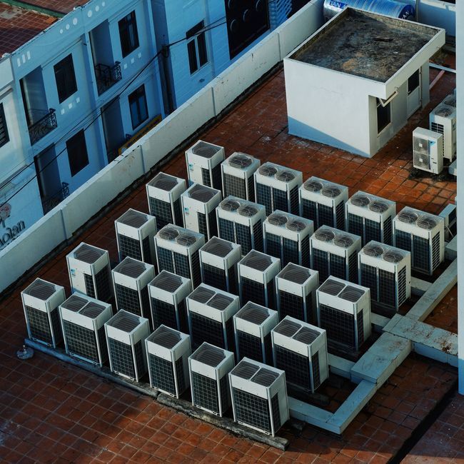 Imagem de uma série de unidades de ar-condicionado instaladas em um telhado de edifício urbano, representando infraestrutura de climatização empresarial.