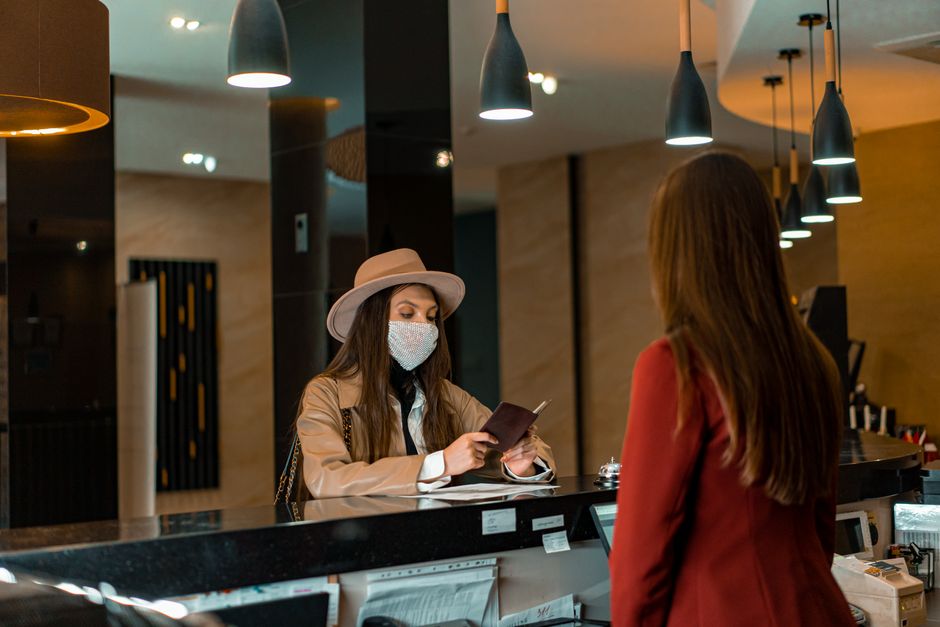 Imagem de uma mulher com máscara facial usando um chapéu e vestimenta elegante interagindo com uma atendente em um espaço de recepção ou bar, ilustrando hospitalidade ou atendimento ao cliente.