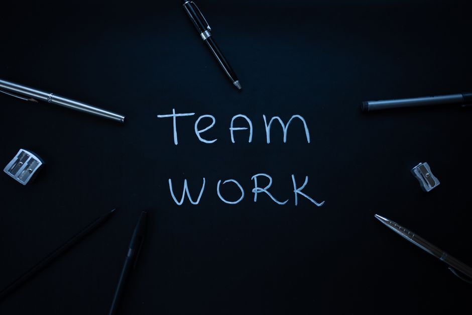 Imagem de uma superfície escura com a frase 'team work' escrita em giz, cercada por canetas, lápis, e itens escolares, simbolizando colaboração e trabalho em equipe.