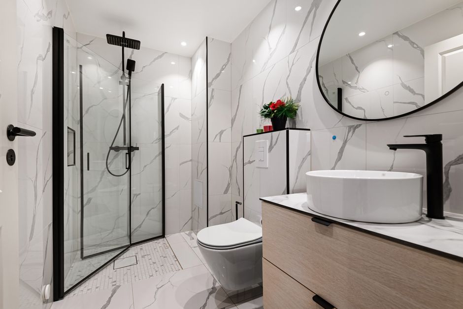 Banheiro moderno com acabamento em mármore, revestido de azulejos brancos com veios cinza, incluindo pia com vaso sanitário e box de vidro fumê, decorado com uma plantinha e espelho redondo.