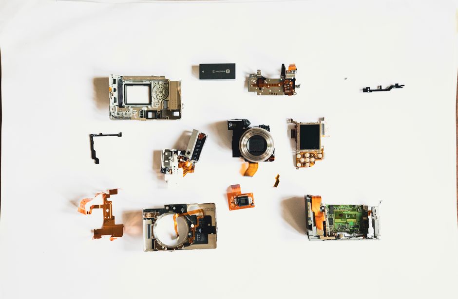 Peças de câmeras digitais desmontadas dispostas sobre uma superfície branca, destacando o processo de reparo ou montagem de tecnologia fotográfica.