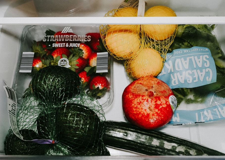 Imagem de uma geladeira com várias frutas frescas, incluindo morangos, melões, pêssegos, uma melancia e pepino, representando alimentação saudável.