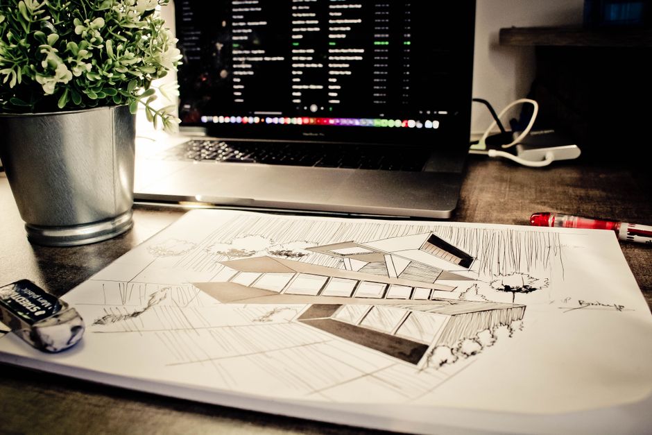 Imagem de uma bancada de trabalho com plantas, laptop com tela de código, e plano de arquitetura, sugerindo foco em tecnologia e planejamento de projetos.