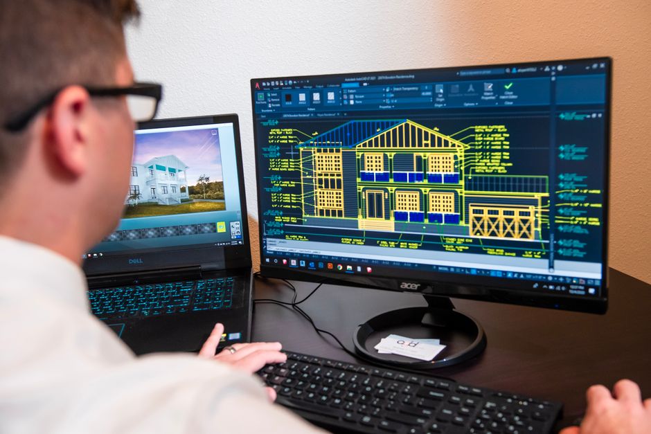 Profissional de arquitetura revisando um projeto detalhado de construção em dois monitores, focado em planejamento e design de edificações