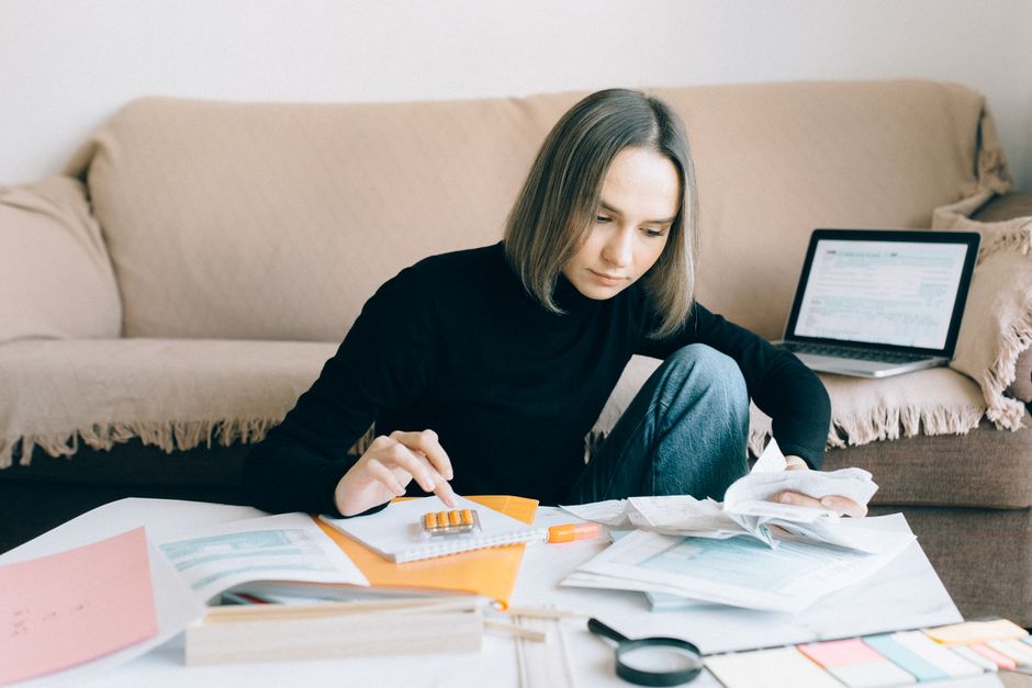Mulher analisando documentos financeiros enquanto trabalha em casa, com computador aberto ao fundo, simbolizando organização e gestão financeira.