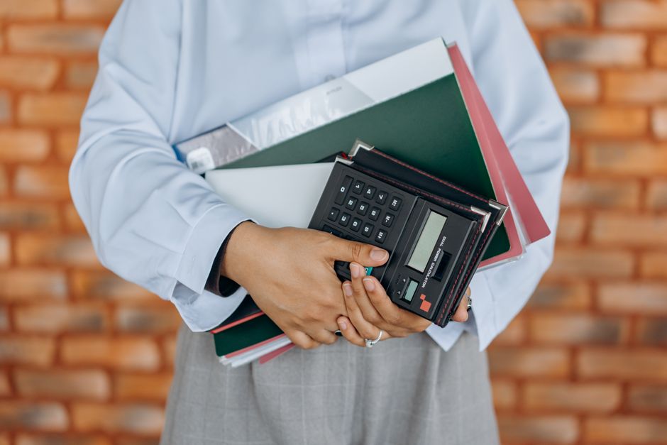 Pessoa segurando materiais de escritório, calculadora e pastas, simbolizando planejamento financeiro ou organização empresarial.