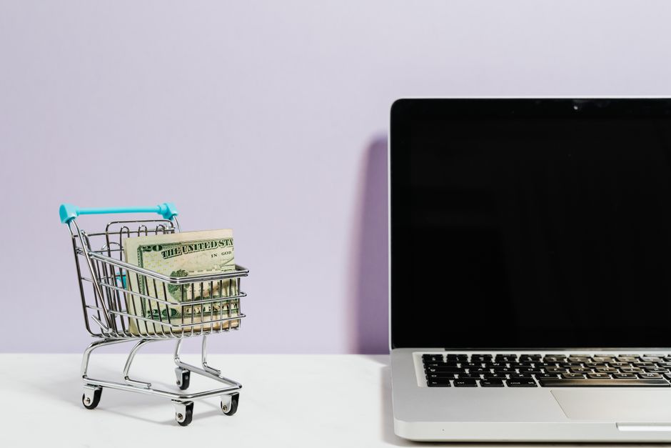 Imagem ilustrativa sobre comércio eletrônico e gestão financeira, mostrando uma miniatura de carrinho de compras com dinheiro dentro ao lado de um laptop.