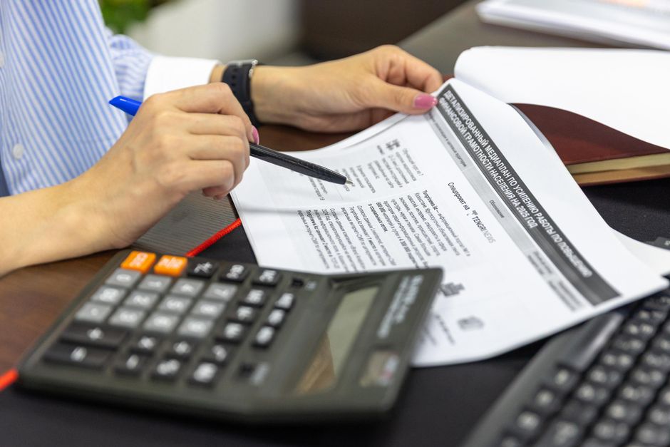 Pessoa analisando documentos financeiros com calculadora e computador em ambiente de escritório, focado em tarefas administrativas e financeiras.