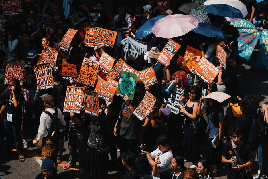 Grupo de manifestantes participando de uma protesto com cartazes e guarda-chuvas na rua, simbolizando mobilização e ativismo social.