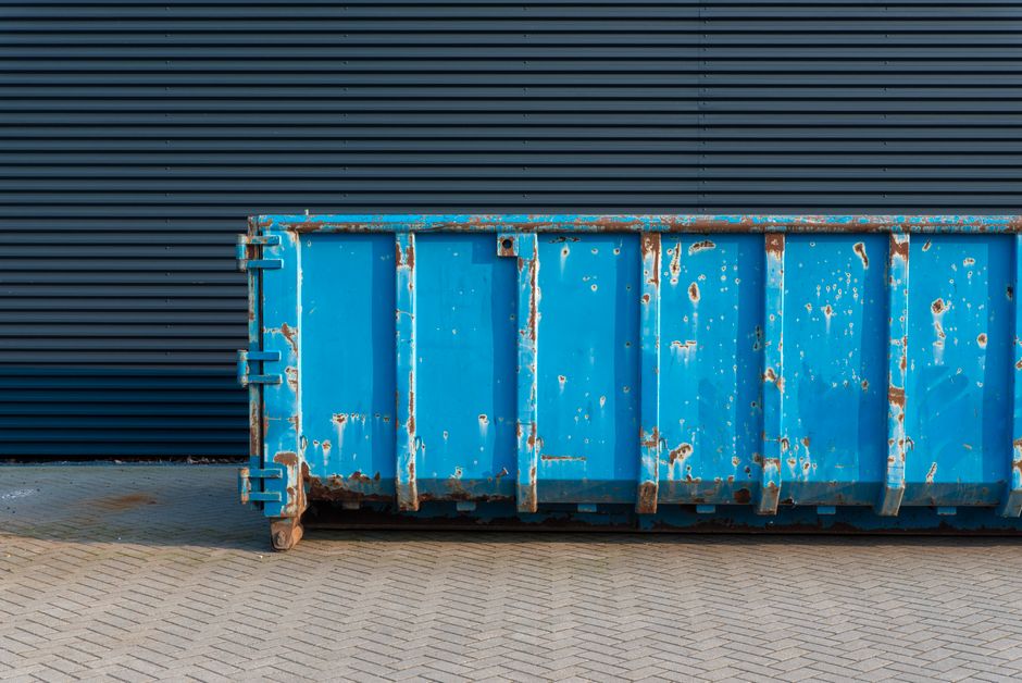 Imagem de um container de lixo azul envelhecido, posicionado em frente a uma parede de metal com linhas horizontais, em um ambiente urbano industrial.