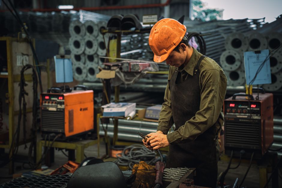 Profissional de soldagem em uma oficina industrial, manipulando equipamentos de solda em um ambiente com ferramentas e materiais metálicos ao fundo.