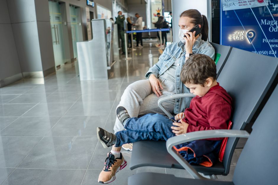 Mulher e criança em uma sala de espera de aeroporto, aguardando o embarque, com ela usando máscara e falando ao telefone enquanto a criança brinca com um dispositivo eletrônico.