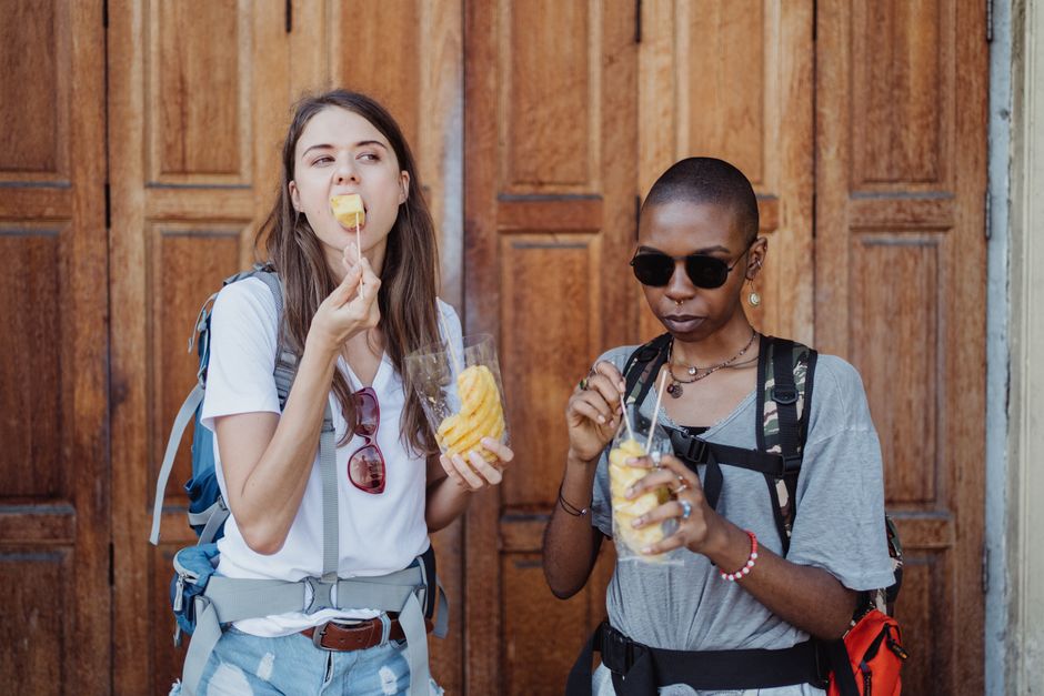 Duas mulheres jovens com mochilas desfrutando de sorvetes ao ar livre, num cenário urbano com fundo de portas de madeira.