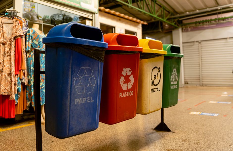 Imagem de lixeiras de cores diferentes em um espaço de reciclagem, destacando a separação de resíduos sólidos.