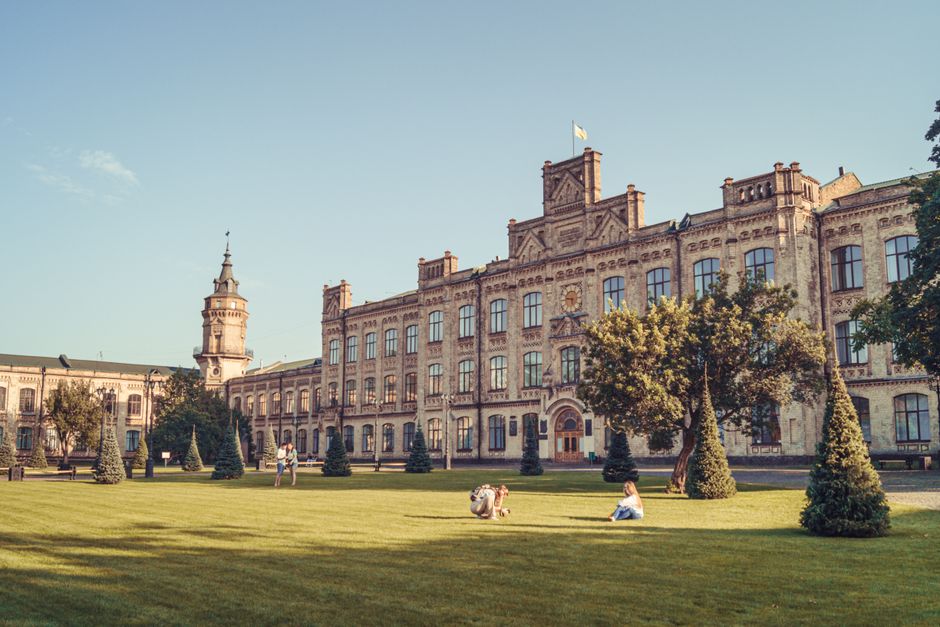 Imagem de um grande edifício histórico com arquitetura clássica, rodeado por um jardim bem cuidado, em um dia ensolarado com poucas pessoas relaxando ao ar livre.