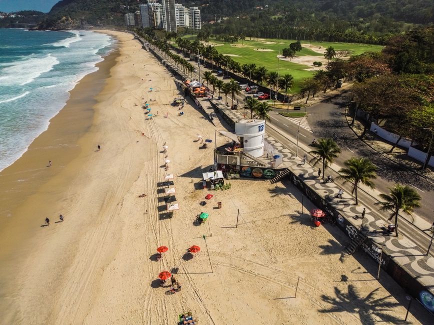Vista aérea de uma praia urbana com área de lazer, calçadão com palm trees e instalação de quiosques.