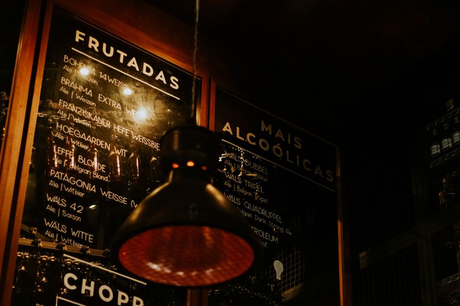 Imagem de um bar à noite com um menu de bebidas ao fundo e uma luminária pendurada em primeiro plano, criando um ambiente urbano e descontraído.