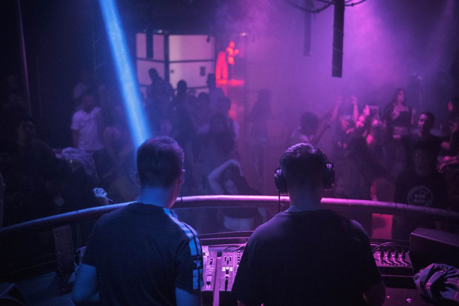 DJs em apresentação ao vivo em uma noite de balada com iluminação colorida e multidão dançando ao fundo.