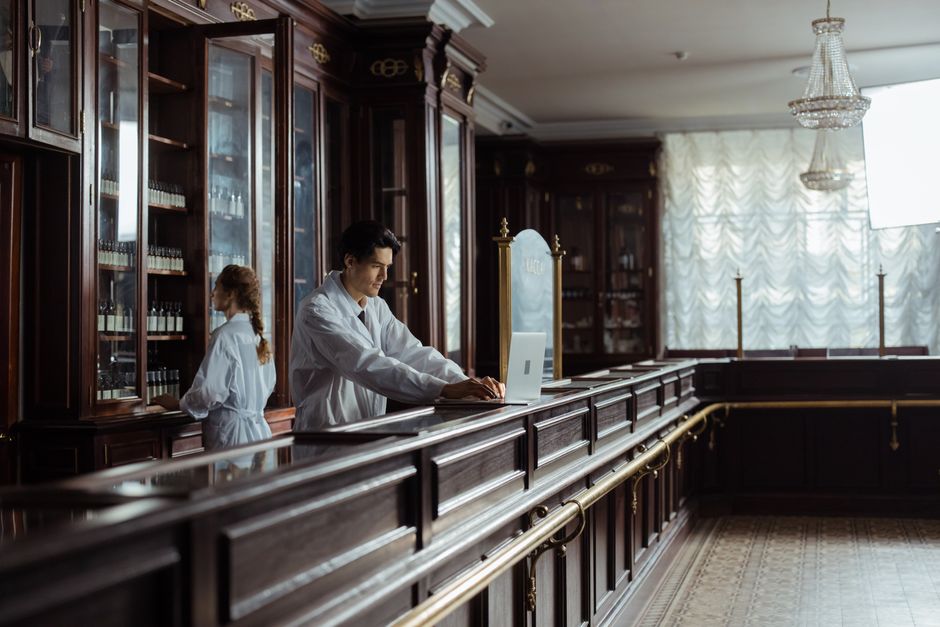 Imagem de um atendente de hotel ou recepcionista futurista usando um laptop atrás do balcão de atendimento em um ambiente clássico com móveis de madeira e cortinas de pano leve ao fundo.
