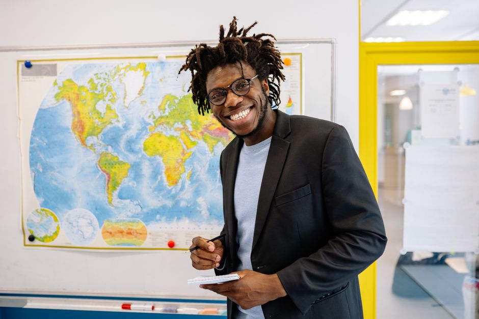 Homem sorridente em ambiente de trabalho escolar ou acadêmico, diante de um mapa mundi na parede.