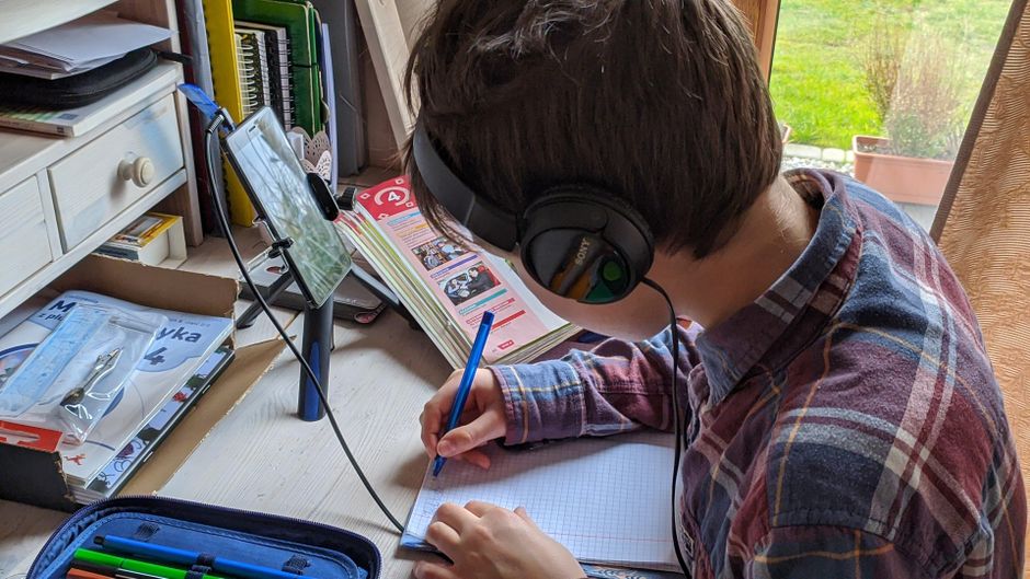 Jovem estudando em casa com fone de ouvido, observando conteúdo digital e escrevendo em caderno