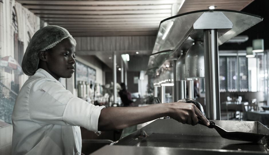 Imagem de uma cozinheira preparando comida em uma cozinha de restaurante, refletindo um ambiente de alimentos e hospitalidade.