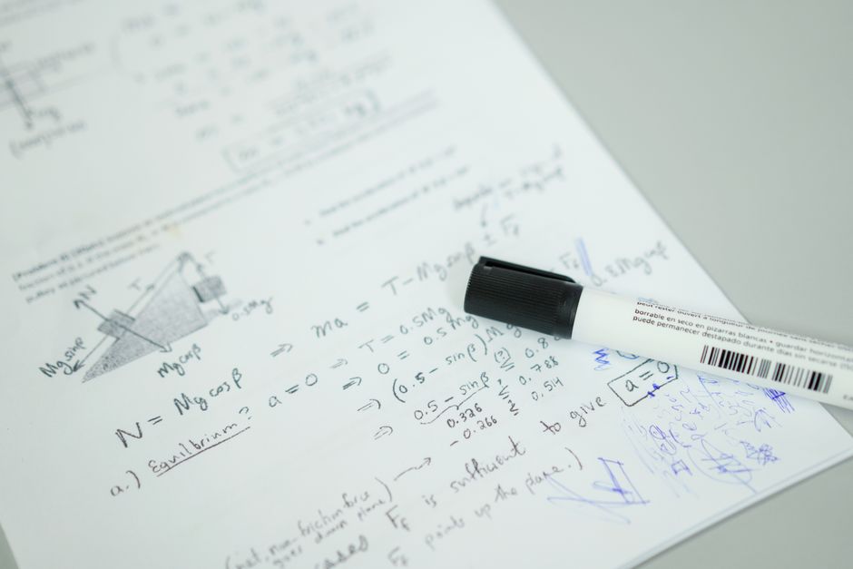 Imagem de uma folha de papel com anotações e fórmulas matemáticas relacionadas à física, com um marcador de quadro branco ao lado, utilizada em artigos sobre estudos acadêmicos ou ensino de ciências.