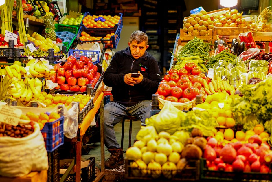 Homem analisando seu telefone enquanto está rodeado de diversas frutas frescas em uma feira ou mercado ao ar livre.