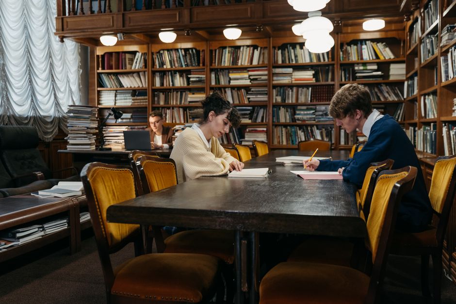 Estudantes em uma sala de estudos com uma biblioteca de fundo, focados em suas tarefas acadêmicas ao lado de um professor ou colega