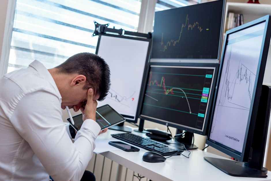 Pessoa frustrada diante de múltiplos monitores exibindo gráficos financeiros, simbolizando a frustração com investimentos ou negociações difíceis.