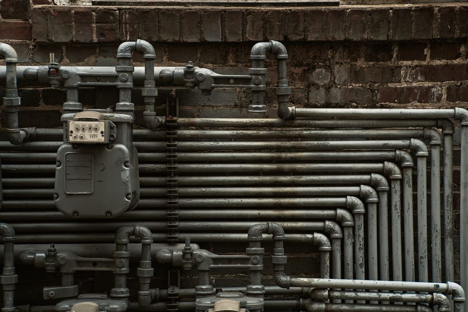 Sistema de encanamento de água ou gás instalado em uma parede de tijolos, demonstrando infraestrutura hidráulica industrial ou residencial.