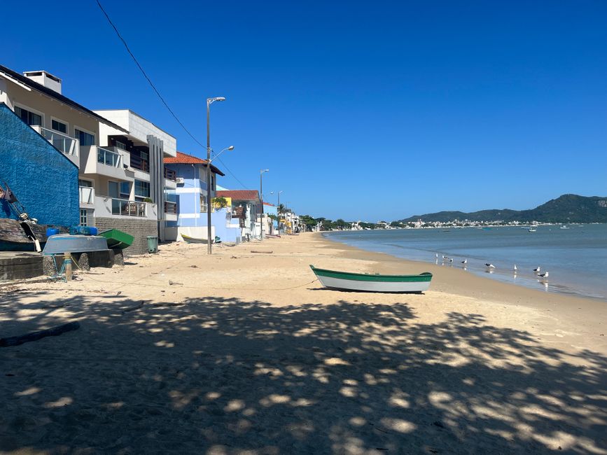 Praia tranquila com casas à beira-mar e um barco deitado na areia, sob um céu azul claro e águas calmas.