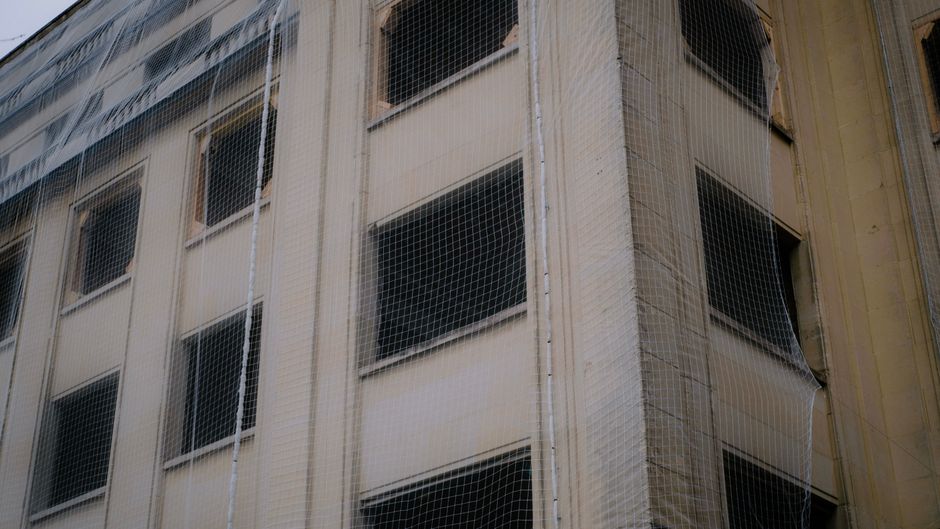 Imagem de um edifício residencial em construção, cercado por redes de proteção e andaimes na estrutura externa, destacando obras de manutenção ou renovação.